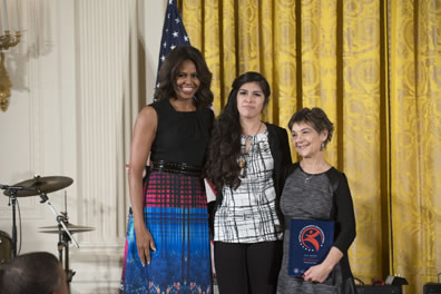 Michelle Obama in black tank top and red, blue dress with checked pattern and flowers, Jennifer MacCaskey in white with black cross patterend fblouse and black sweater and pants and cross necklace, Halperin in grey dress with black sleeves holding award. American flag behind first lady, gold curtain across the back to the right, drum set to the left.